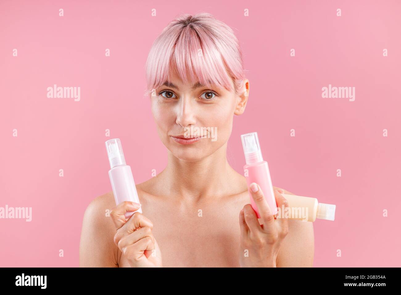Attraktive junge Frau mit rosa Haaren, die die Kamera anschaut, drei Reisetaschen aus Kunststoff mit Beauty-Produkten in der Hand hält und isoliert über Rosa posiert Stockfoto