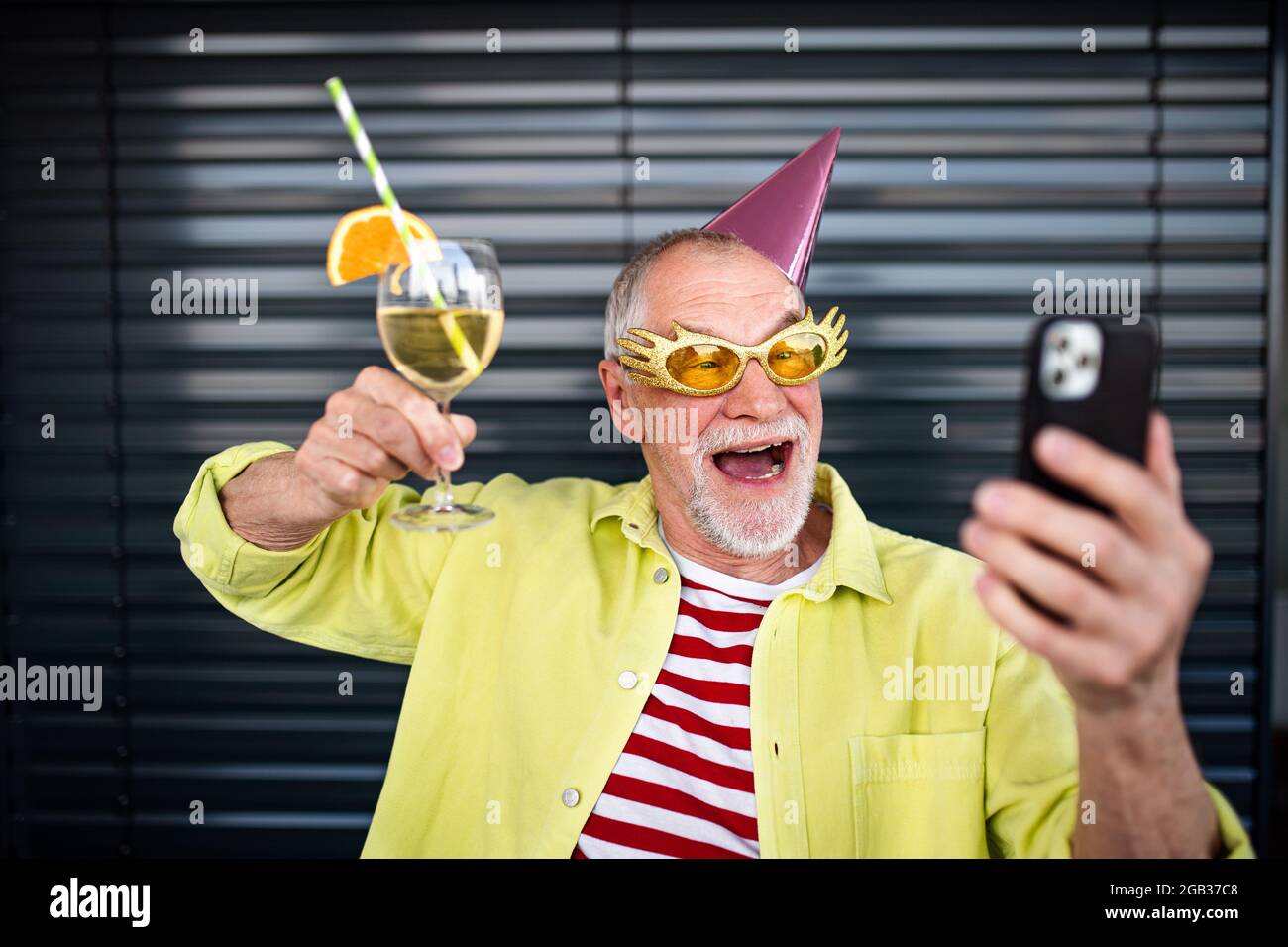 Porträt eines älteren Mannes mit Partyhut und Getränk, der im Freien steht und Selfie macht. Stockfoto