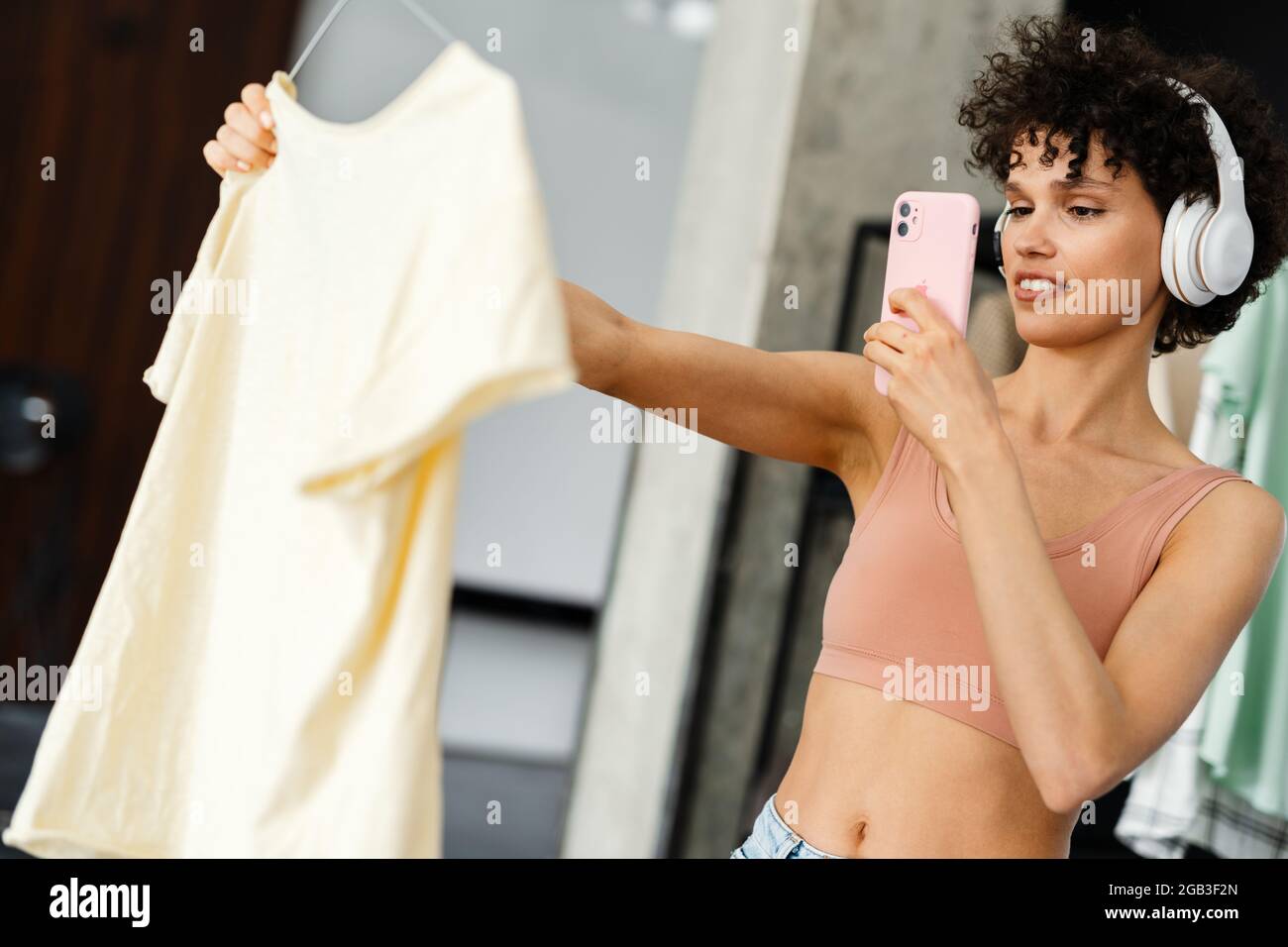 Lächelnde junge Frau mit Kopfhörern, die ein Foto von einem T-Shirt gemacht hat, das zu Hause das richtige Kleidungsstück ausgewählt hat Stockfoto