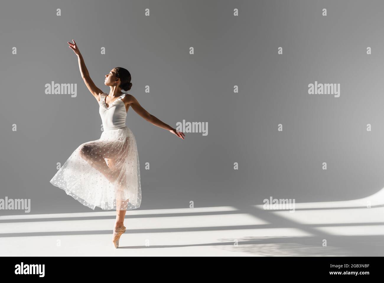 Seitenansicht der Ballerina in weißem Rock, die auf einem Bein auf grauem Hintergrund mit Sonnenlicht steht Stockfoto
