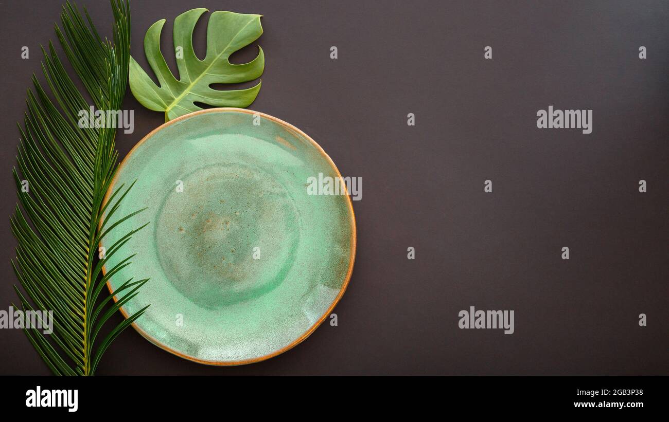 Leere grüne Platte auf schwarzem Hintergrund. Luxuriöser Speiseteller, serviert mit einer Monstera-Pflanze im asiatischen Kinfolk-Grunge-Stil mit tropischen Palmenblättern. Modell leer Stockfoto