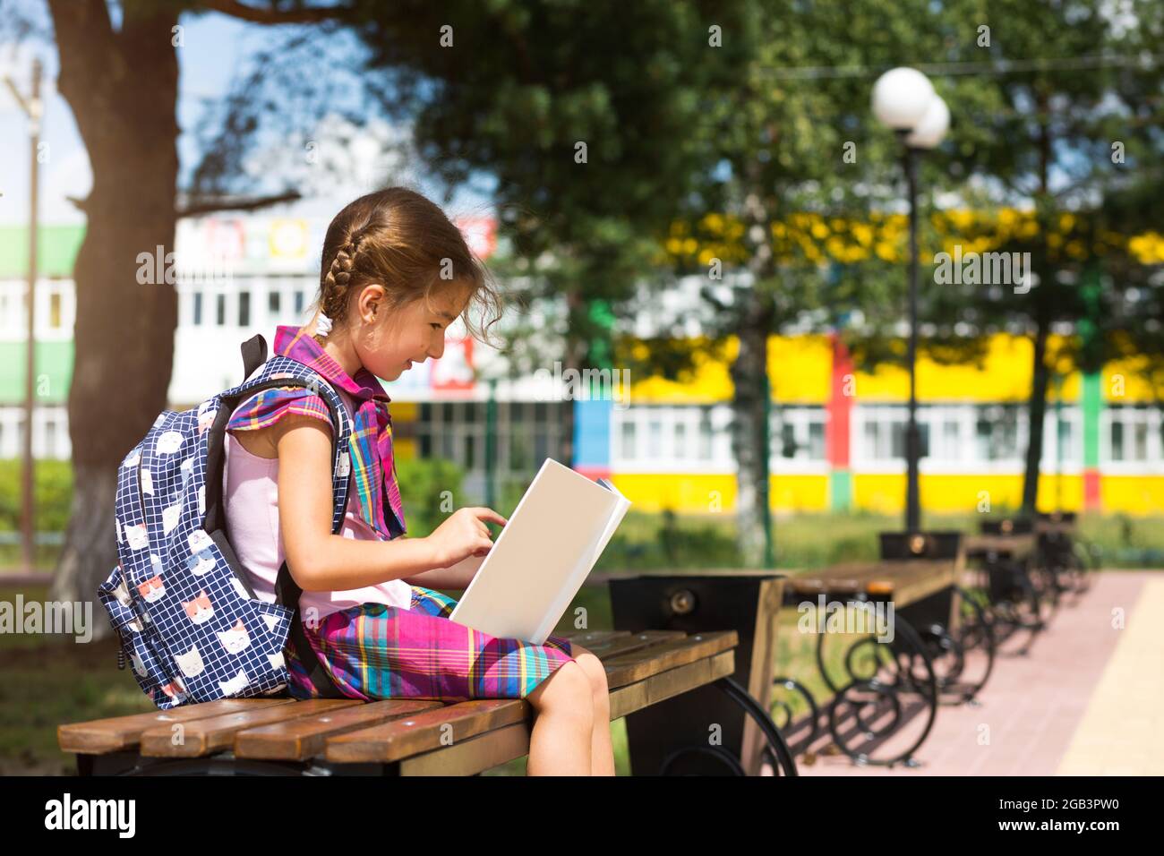 Mädchen mit Rucksack, das auf einer Bank sitzt und ein Buch in der Nähe der Schule liest. Zurück zur Schule, Stundenplan, ein Tagebuch mit Noten. Bildung, Grundschule en Stockfoto