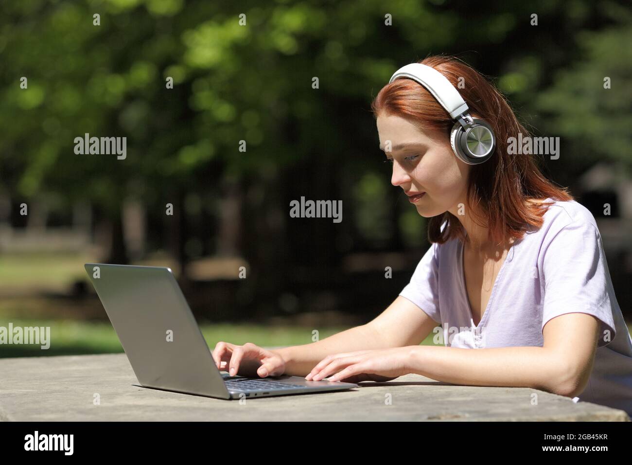 Profil einer Frau mit kabellosen Kopfhörern, die ihren Laptop in einem Park überprüft Stockfoto