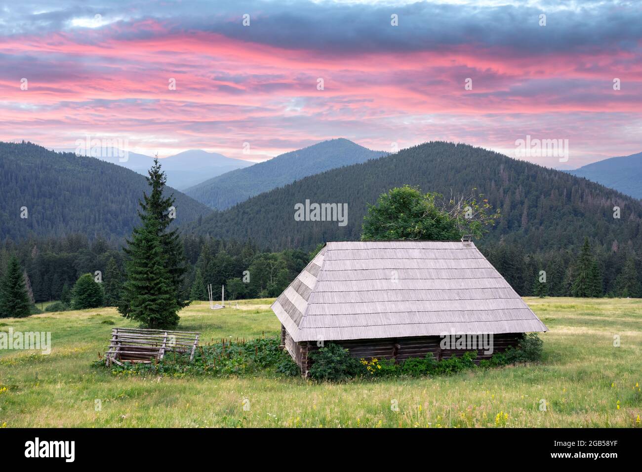 Malerische Sommerwiese mit altem Holzhaus und purpurem Sonnenuntergangshimmel in den Karpaten, Ukraine. Landschaftsfotografie Stockfoto