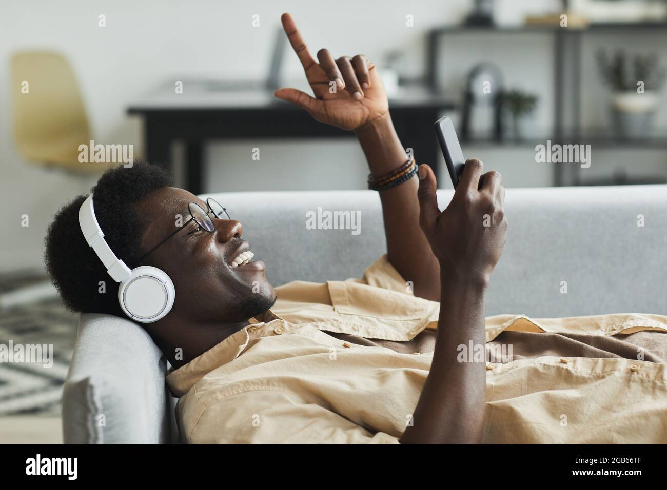 Afrikanischer Mann, der Kopfhörer und Mobiltelefon für Online-Gespräche verwendet, während er auf dem Sofa im Zimmer liegt Stockfoto