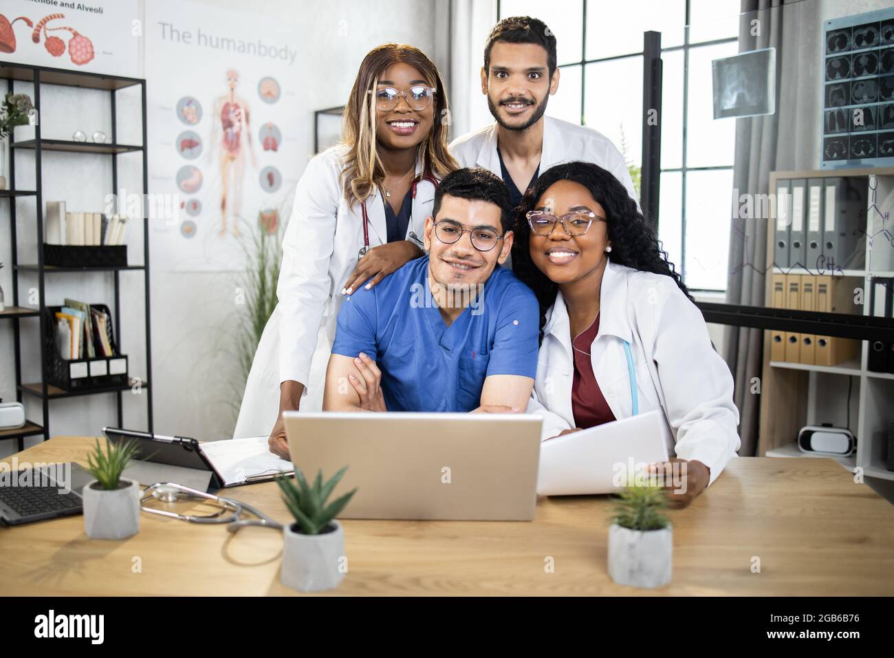 Gruppe von vier internationalen Ärzten in Laborkittel und -Peelings, die zusammen am Tisch sitzen und Laptop und Tablet benutzen. Medizinische Konferenz von männlichen und weiblichen Spezialisten. Mit Wissen teilen. Stockfoto