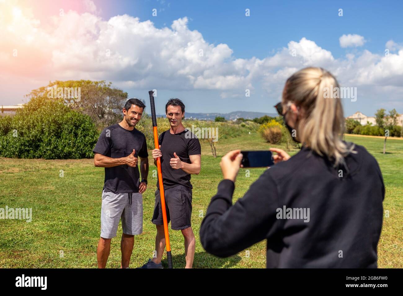 Fitnesstrainer und Klientin posieren für eine Reporterin und schießen auf einem Smartphone. Sonniger Tag. Mit Gymnastikstab. Sommer im Garten. Stockfoto