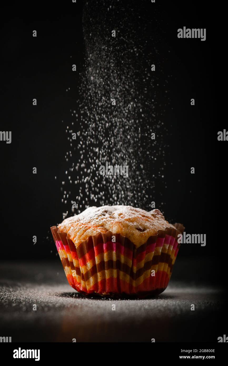 Der Koch streut Muffins mit Puderzucker auf schwarzem Hintergrund. Der Prozess der Herstellung von hausgemachtem Kuchen. Vertikales Foto. Stockfoto
