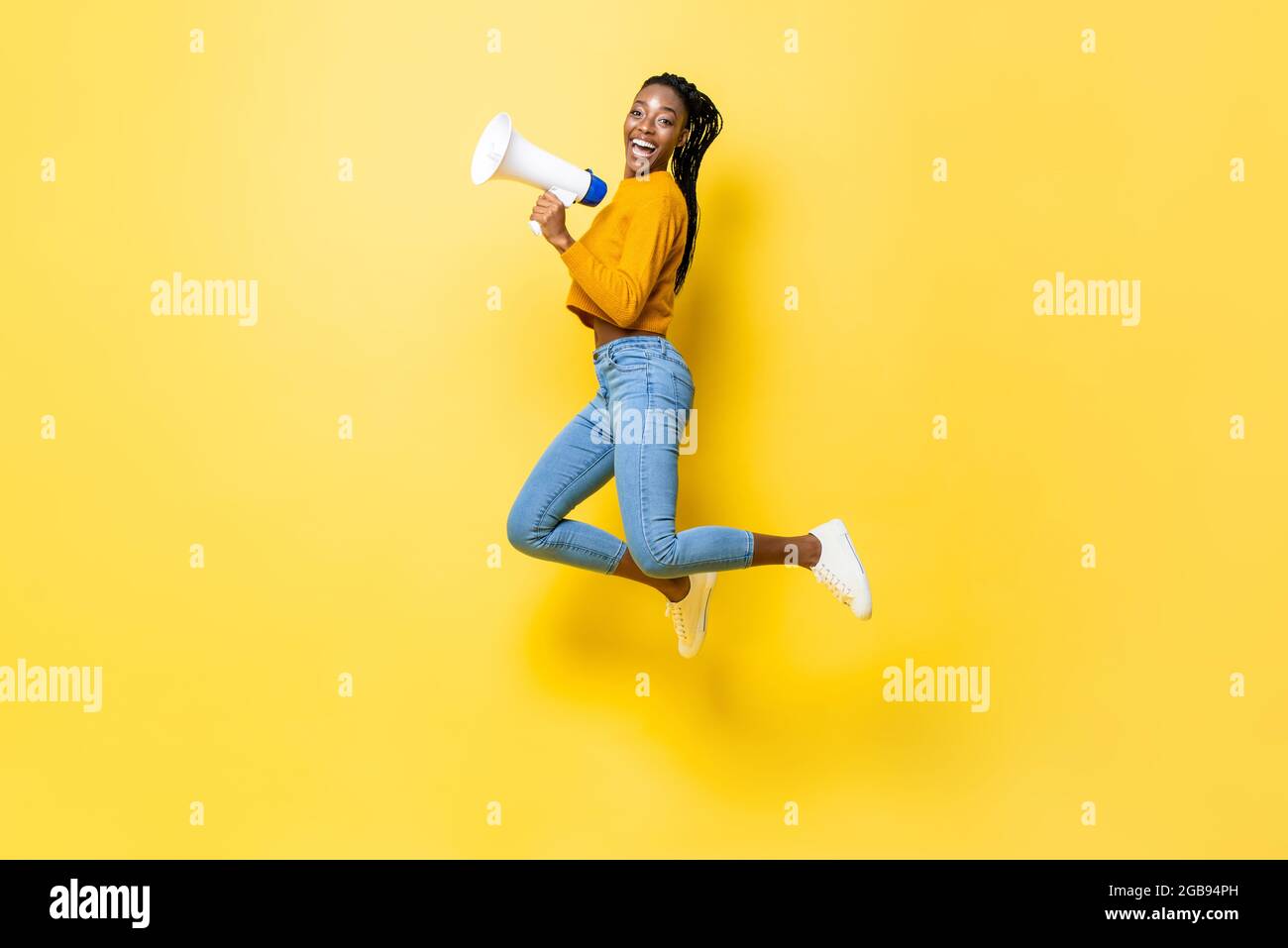 Fröhliche junge afroamerikanische Frau hält Megaphon springen und Ankündigung in isolierten Studio gelben Hintergrund Stockfoto