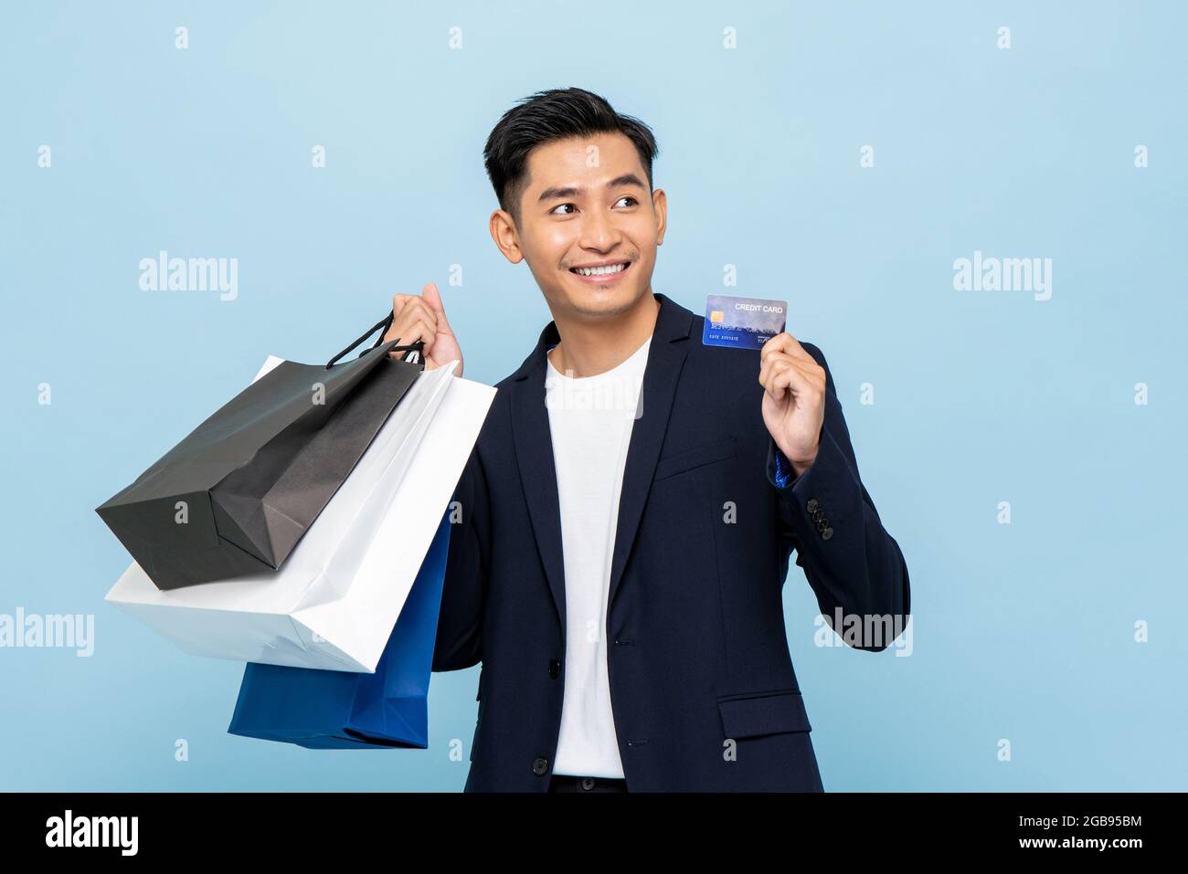 Waist-up-Porträt des jungen schönen asiatischen Mann hält Einkaufstasche und Kreditkarte für bargeldlose Zahlung Konzept in isolierten Studio hellblau backgrou Stockfoto