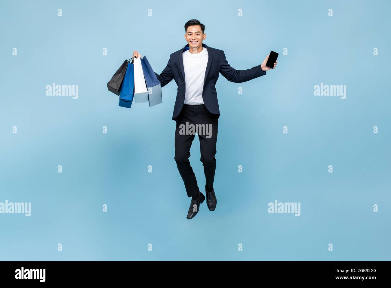 Aufgeregt junge gut aussehende asiatische Mann hält Handy und Einkaufstaschen springen in hellblauen Studio isoliert Hintergrund Stockfoto