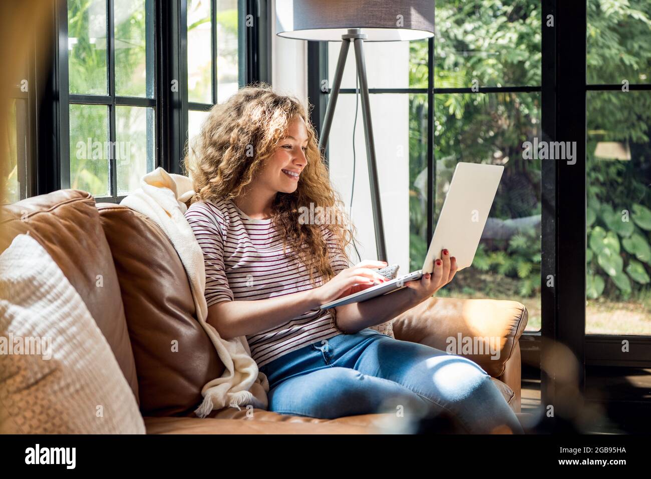 Glücklich lächelnde kaukasische Frau, die tagsüber mit einem Laptop auf der Couch im Wohnzimmer zu Hause im Internet surfte Stockfoto