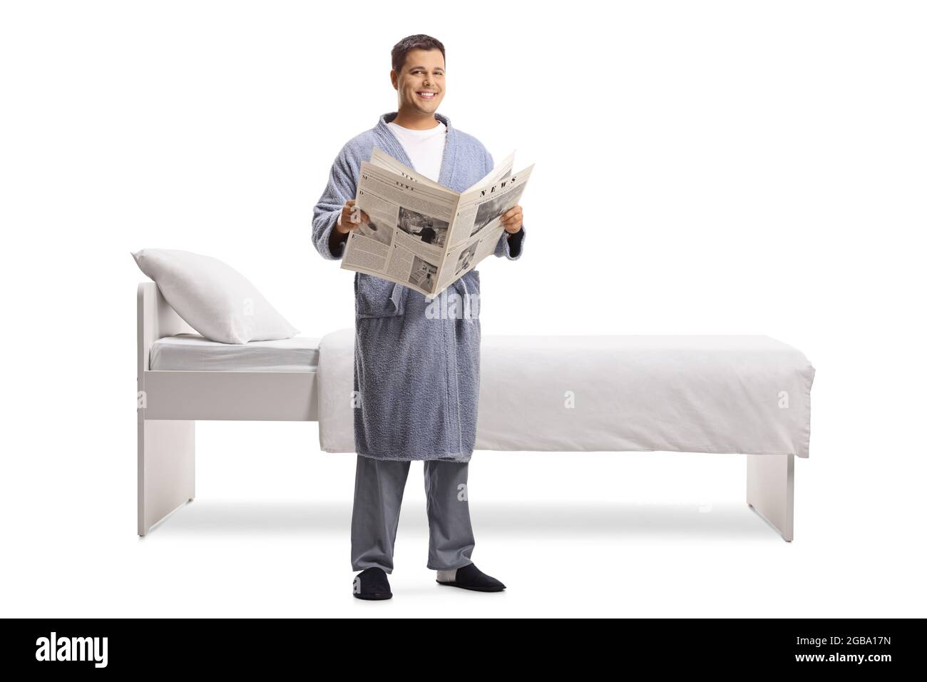 Junger Mann mit einer Zeitung, die einen Bademantel trägt und neben einem Bett auf weißem Hintergrund steht Stockfoto