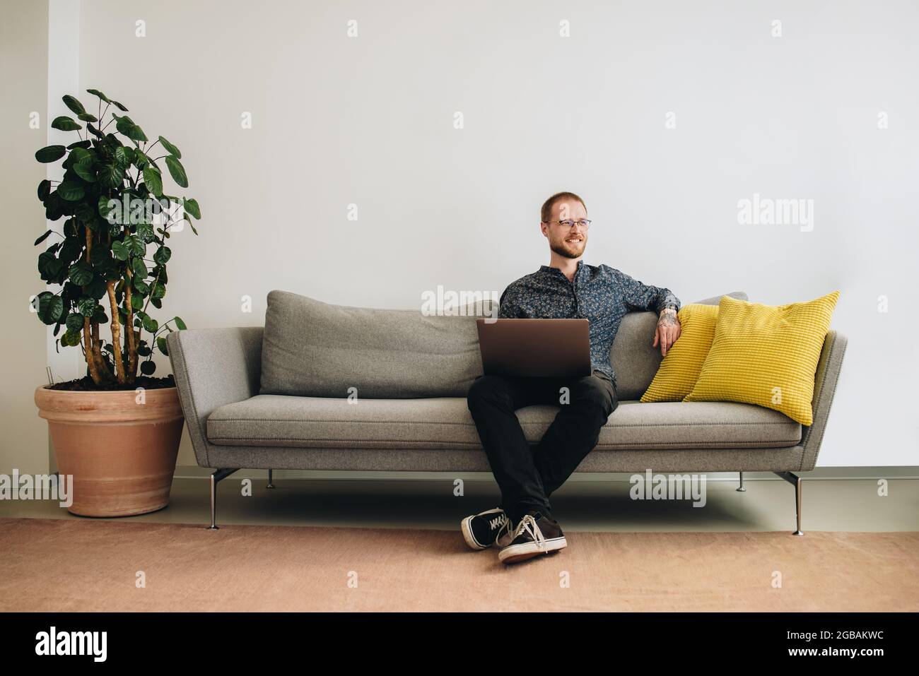 Geschäftsmann mit Laptop auf Sofa. Männlicher Mitarbeiter, der wegschaut, während er in der Bürolobby an einem Laptop arbeitet. Stockfoto