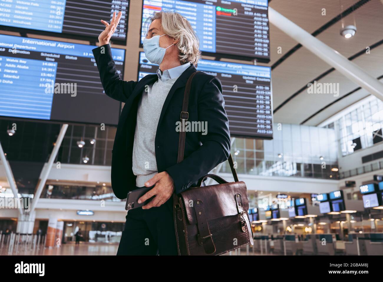 Geschäftsmann mit Gesichtsmaske am Flughafenterminal winkt jemanden an. Geschäftsreisende, die nach der Sperre von Covid-19 reisen. Stockfoto