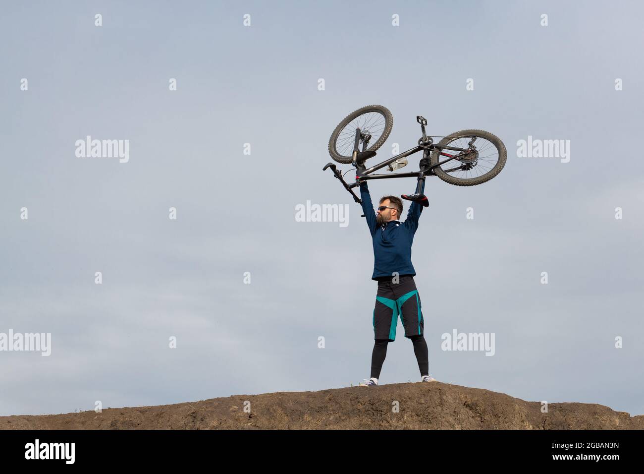 Bärtiger Radfahrer auf dem Berg hält sein Fahrrad in seinen Armen über seinem Kopf Stockfoto