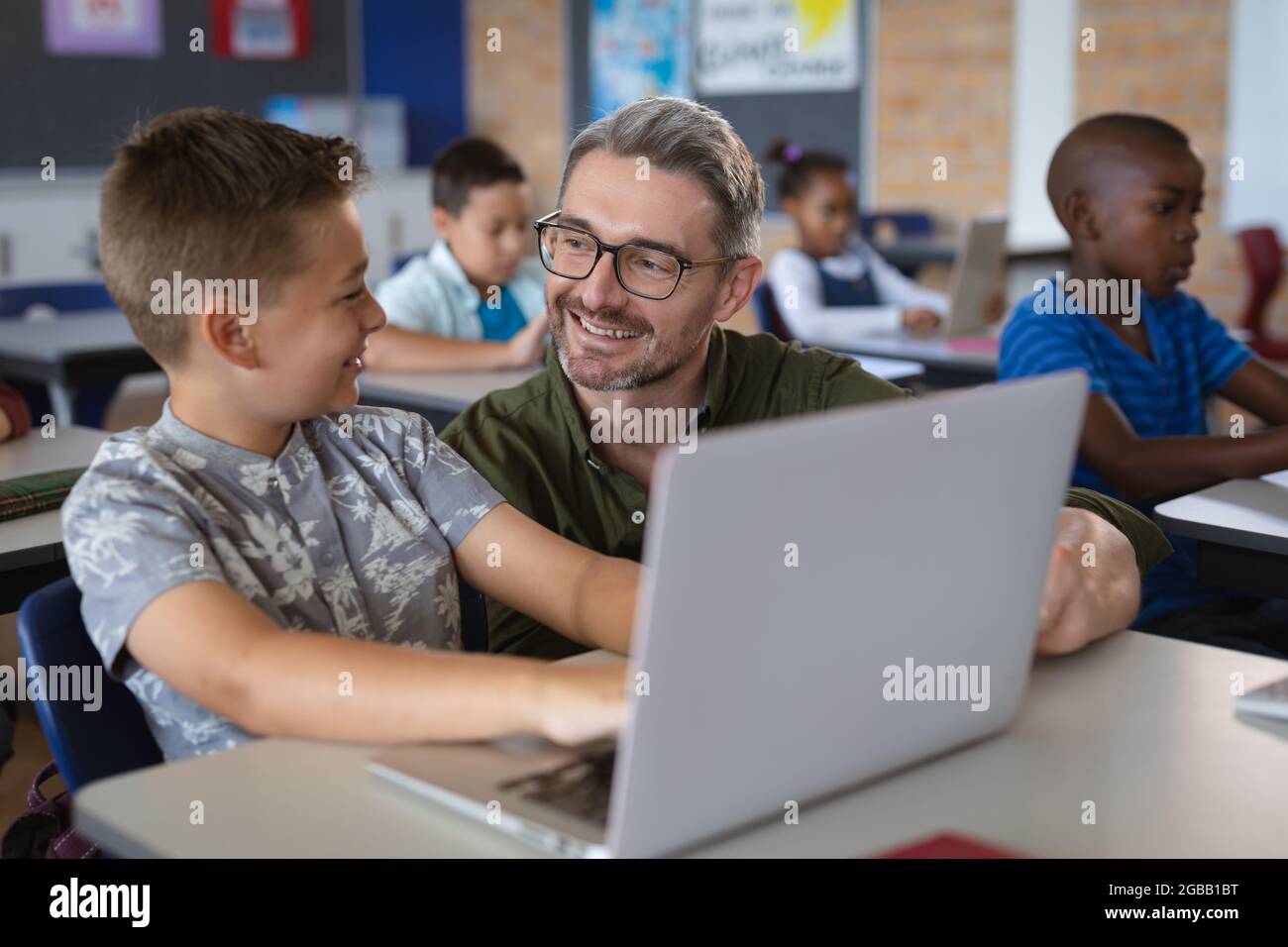 Kaukasischer männlicher Lehrer, der einem kaukasischen Jungen beibringt, wie er in der Schule in der Klasse einen Laptop benutzt Stockfoto