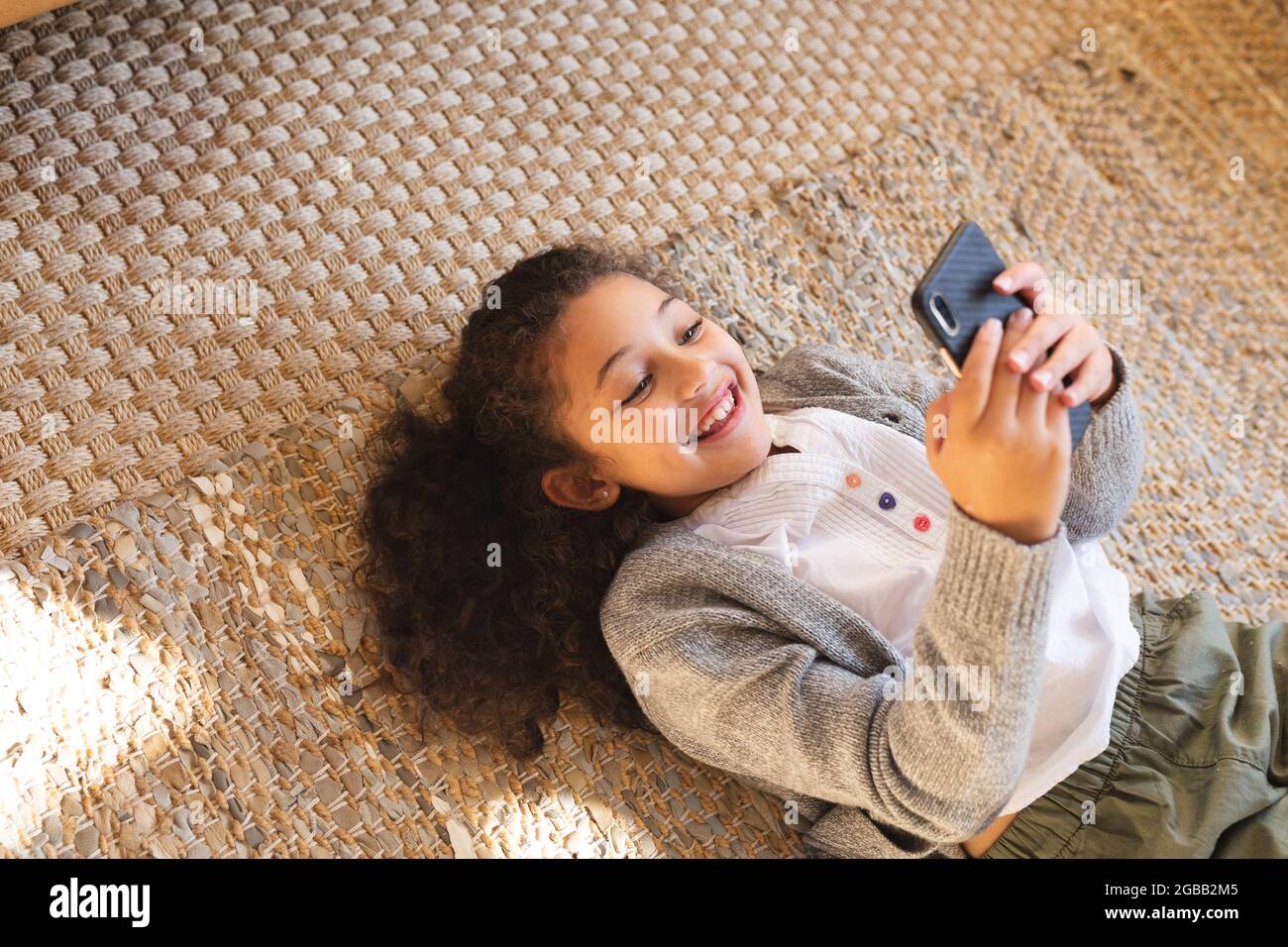 Lächelndes Mädchen mit gemischter Rasse, das mit dem Smartphone auf dem Teppich liegt Stockfoto