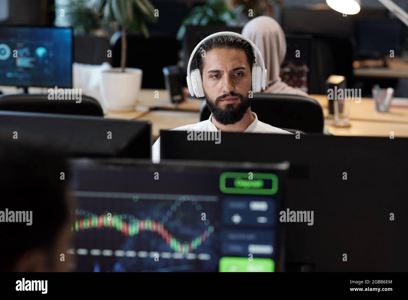 Bärtiger arabischer Geschäftsmann mit Kopfhörern, der in einer Büroumgebung auf den Computerbildschirm schaut Stockfoto
