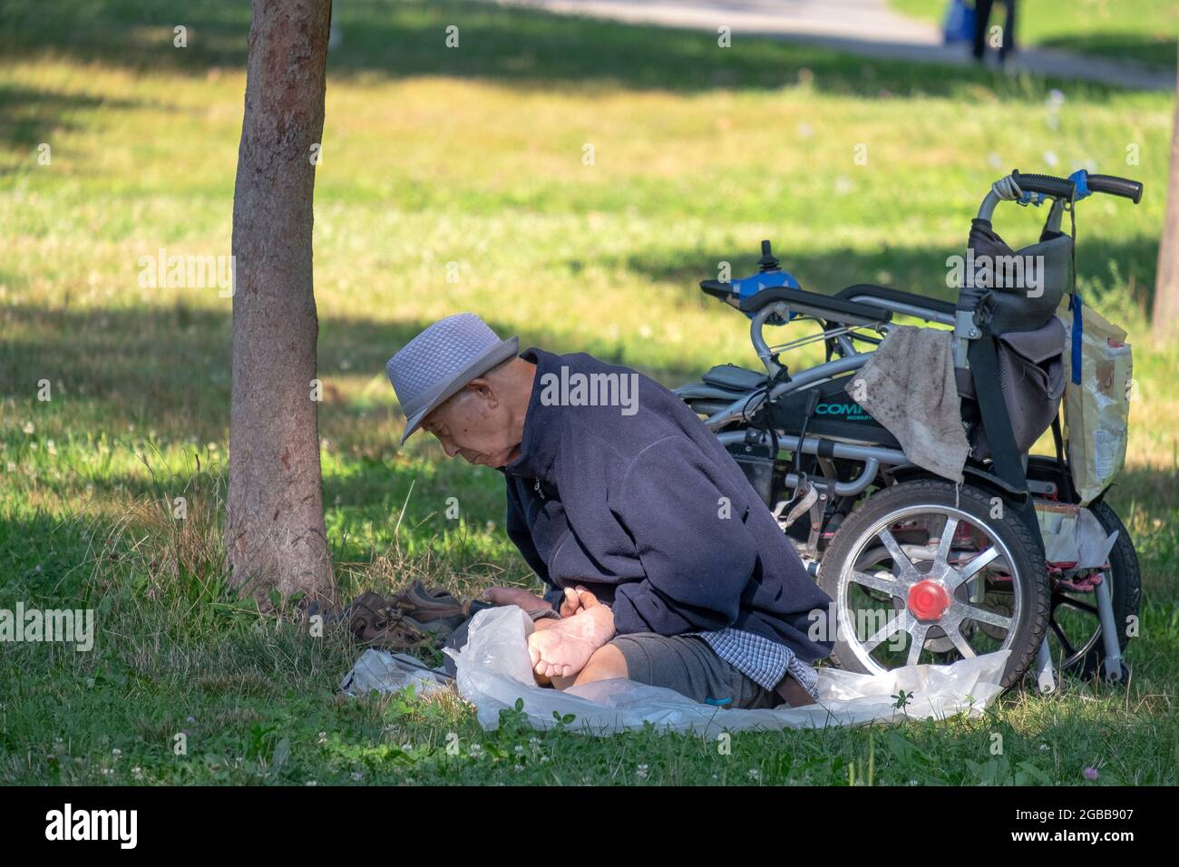 Ein chinesischer Amerikaner in den 80ern macht die langsamen Falun Dafa-Übungen, während er neben einem Baum und in der Nähe seines Rollstuhls sitzt. In einem Park in Queens, New York Stockfoto