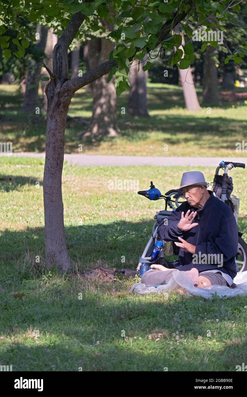 Ein sitzender 86-jähriger Mann macht in der Nähe eines Baumes langsam laufende Falun Gong-Übungen. In einem Park in Queens, New York, ein sehr vielfältiger Ort. Stockfoto