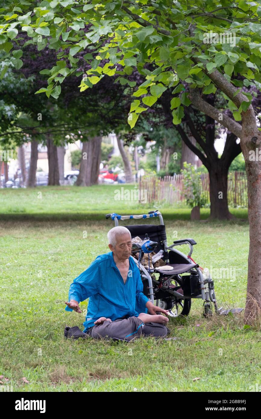 Ein sitzender 86-jähriger Mann macht in der Nähe eines Baumes langsam laufende Falun Gong-Übungen. In einem Park in Queens, New York, ein sehr vielfältiger Ort. Stockfoto