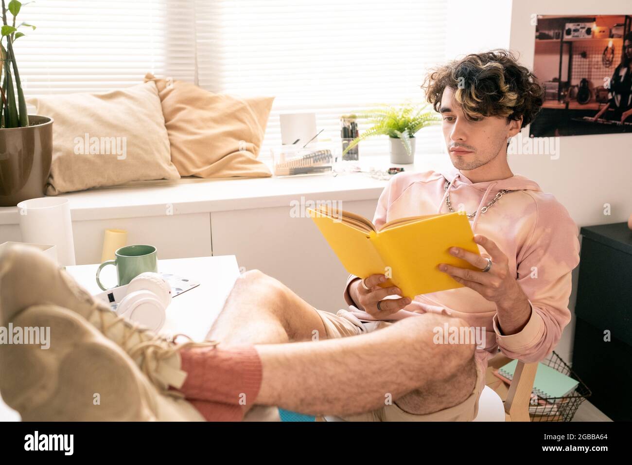 Erholsamer Teenager, der auf einem Stuhl saß und mit seinen Beinen auf dem Schreibtisch ein Buch las Stockfoto