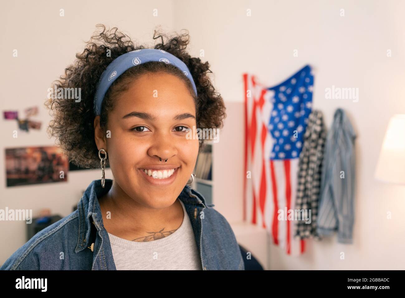 Glückliche afrikanische Teenager in Casualwear, die auf dem Hintergrund einer amerikanischen Flagge in ihrem Zimmer steht Stockfoto