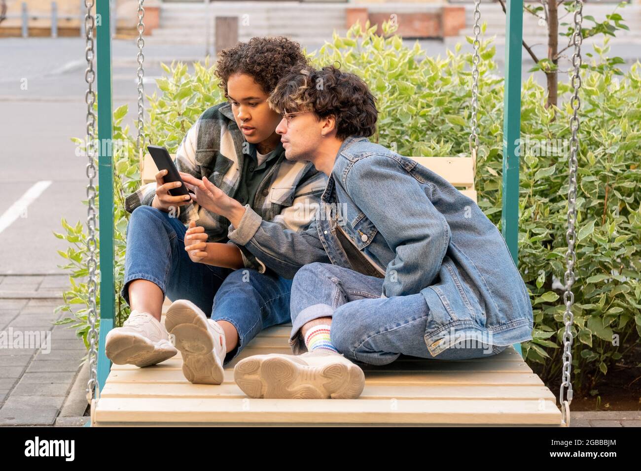 Interkulturelles Teenager-Paar, das auf Schaukeln sitzt und im Smartphone scrollt Stockfoto