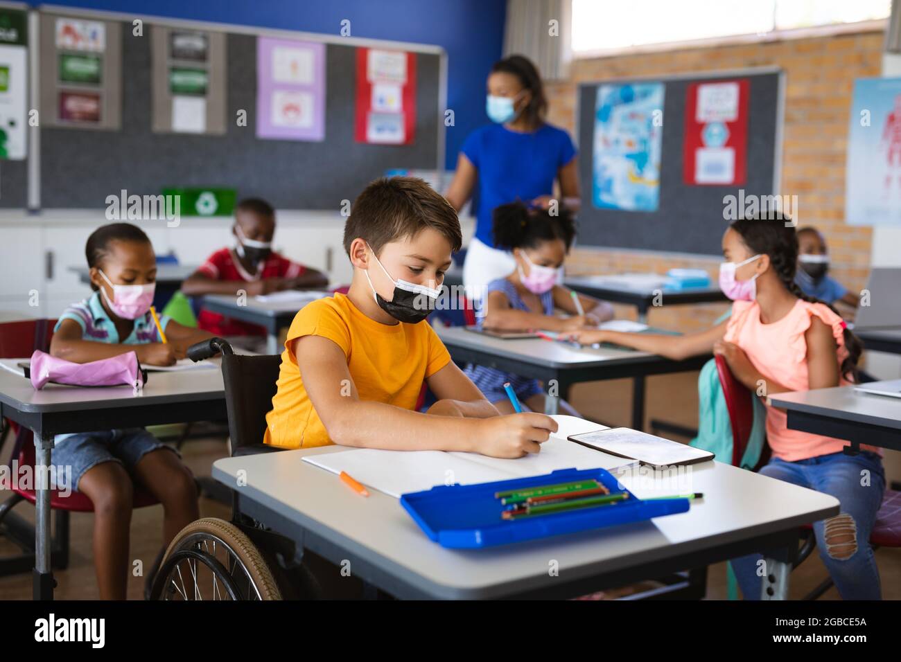 Behinderter kaukasischer Junge mit Gesichtsmaske beim Studieren, während er in der Grundschule im Rollstuhl sitzt Stockfoto
