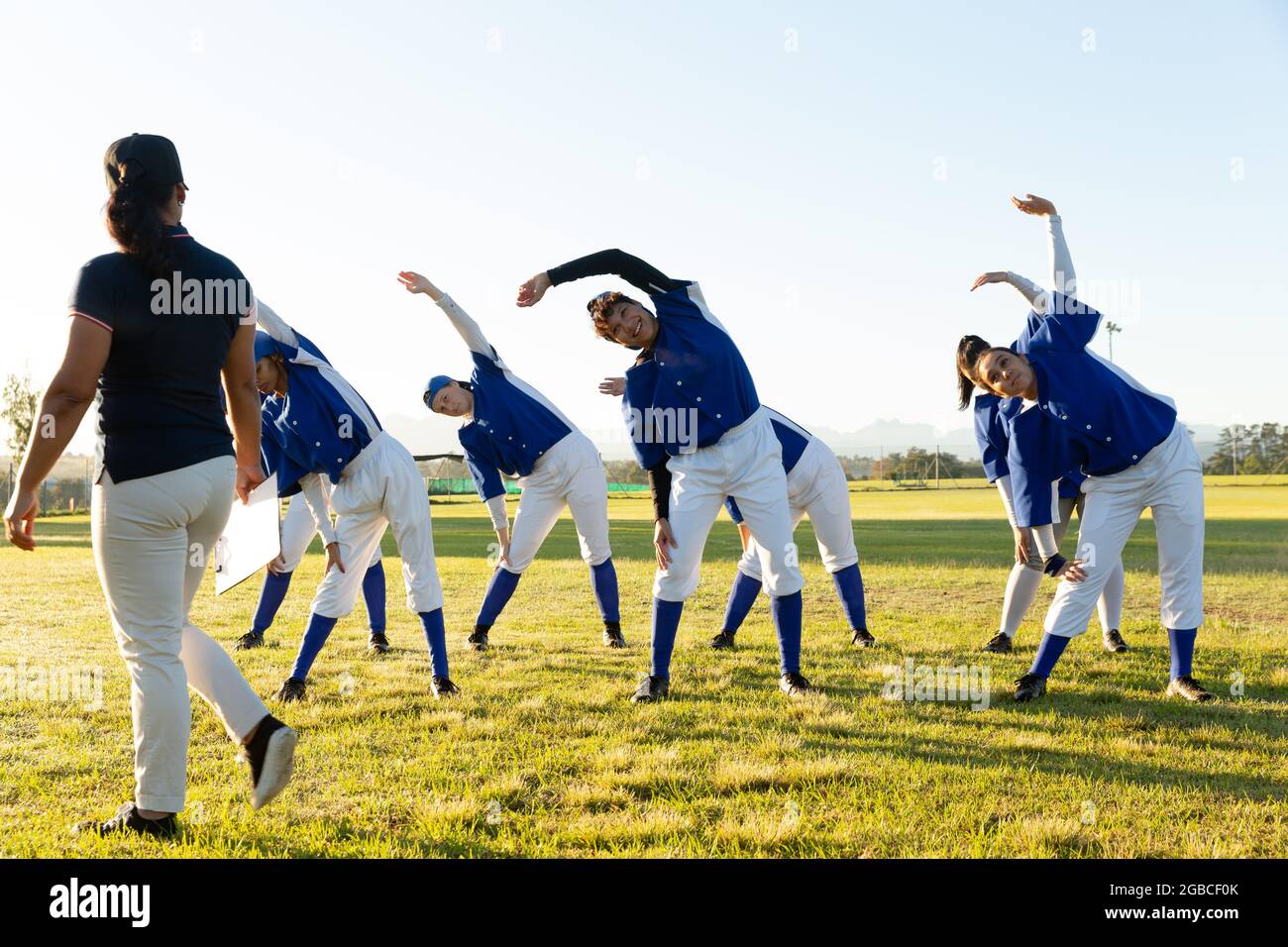Verschiedene Baseballspielerinnen mit Trainer, die sich auf dem Spielfeld aufwärmen und sich von der Hüfte ausstrecken Stockfoto