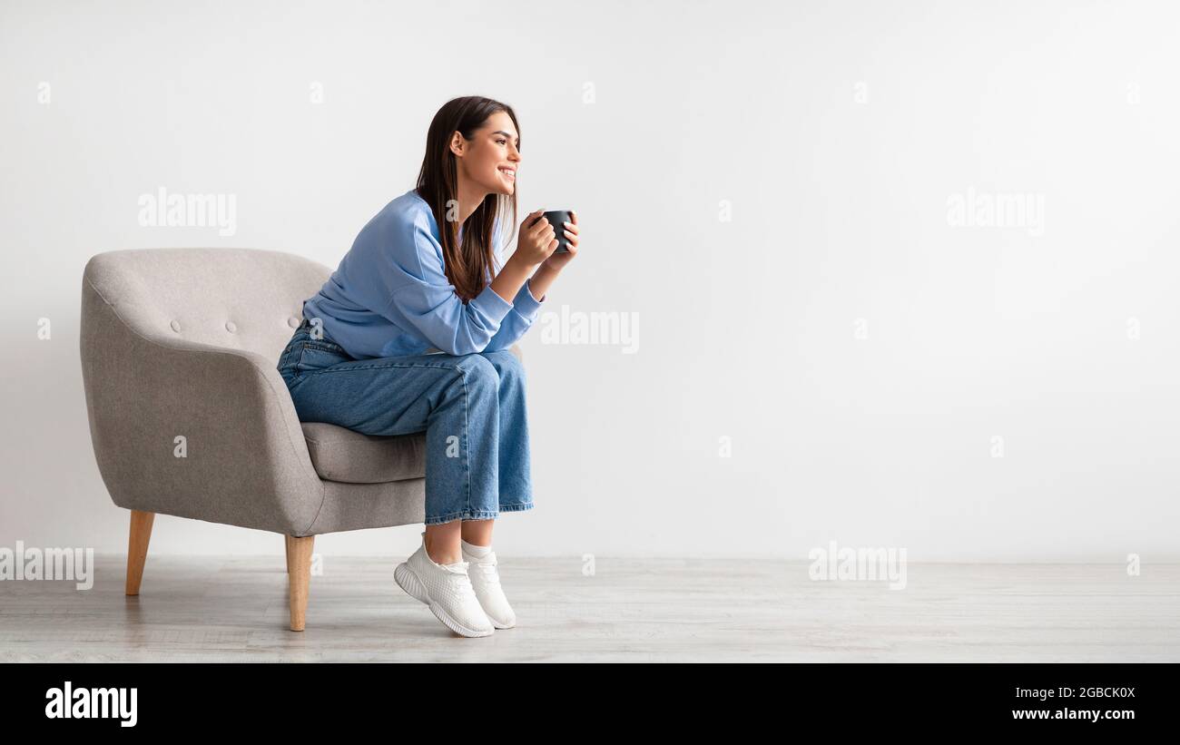 Die ganze Länge der jungen Frau mit Kaffeepause in gemütlichen Sessel gegen weiße Studiowand, Banner mit freiem Platz Stockfoto