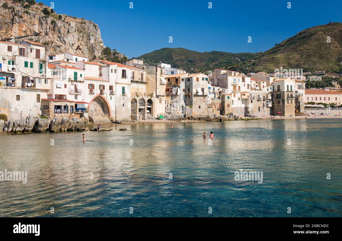 Cefalù, Palermo, Sizilien, Italien. Blick über den ruhigen Hafen auf die Altstadt, überhängende Häuser, die sich entlang der Uferpromenade unter La Rocca gruppieren. Stockfoto
