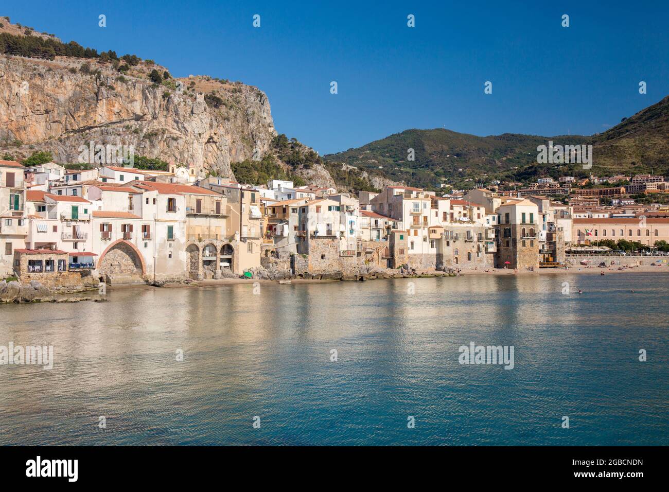 Cefalù, Palermo, Sizilien, Italien. Blick über den ruhigen Hafen auf die Altstadt, überhängende Häuser, die sich entlang der Uferpromenade unter La Rocca gruppieren. Stockfoto