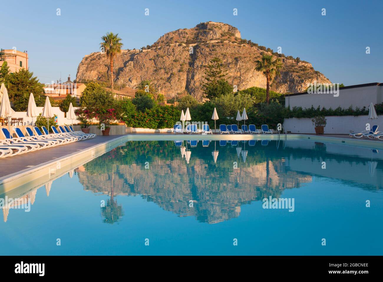 Cefalù, Palermo, Sizilien, Italien. Die hoch aufragenden, sonnendurchfluteten Klippen von La Rocca spiegeln sich im ruhigen Swimmingpool des Hotel Kalura bei Sonnenaufgang wider. Stockfoto