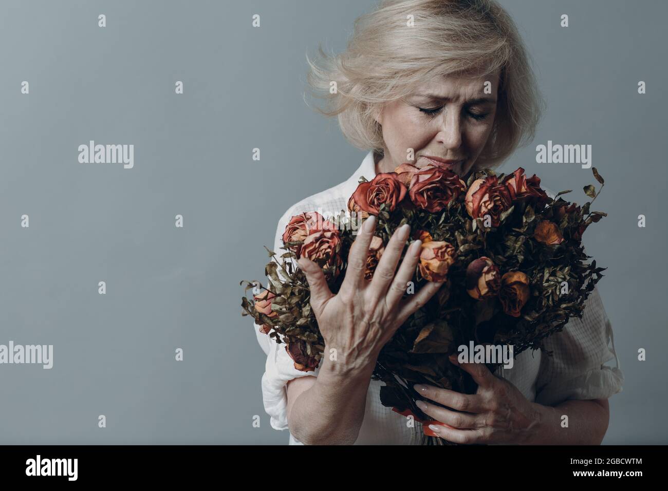 Traurige ältere Frau, die einen Strauß verwelkter Rosenblüten berührt. Grauhaarige reife Dame in Depression. Konzept des alten Alterns. Studio-Aufnahmen auf grauem Hintergrund isoliert. Speicherplatz kopieren Stockfoto