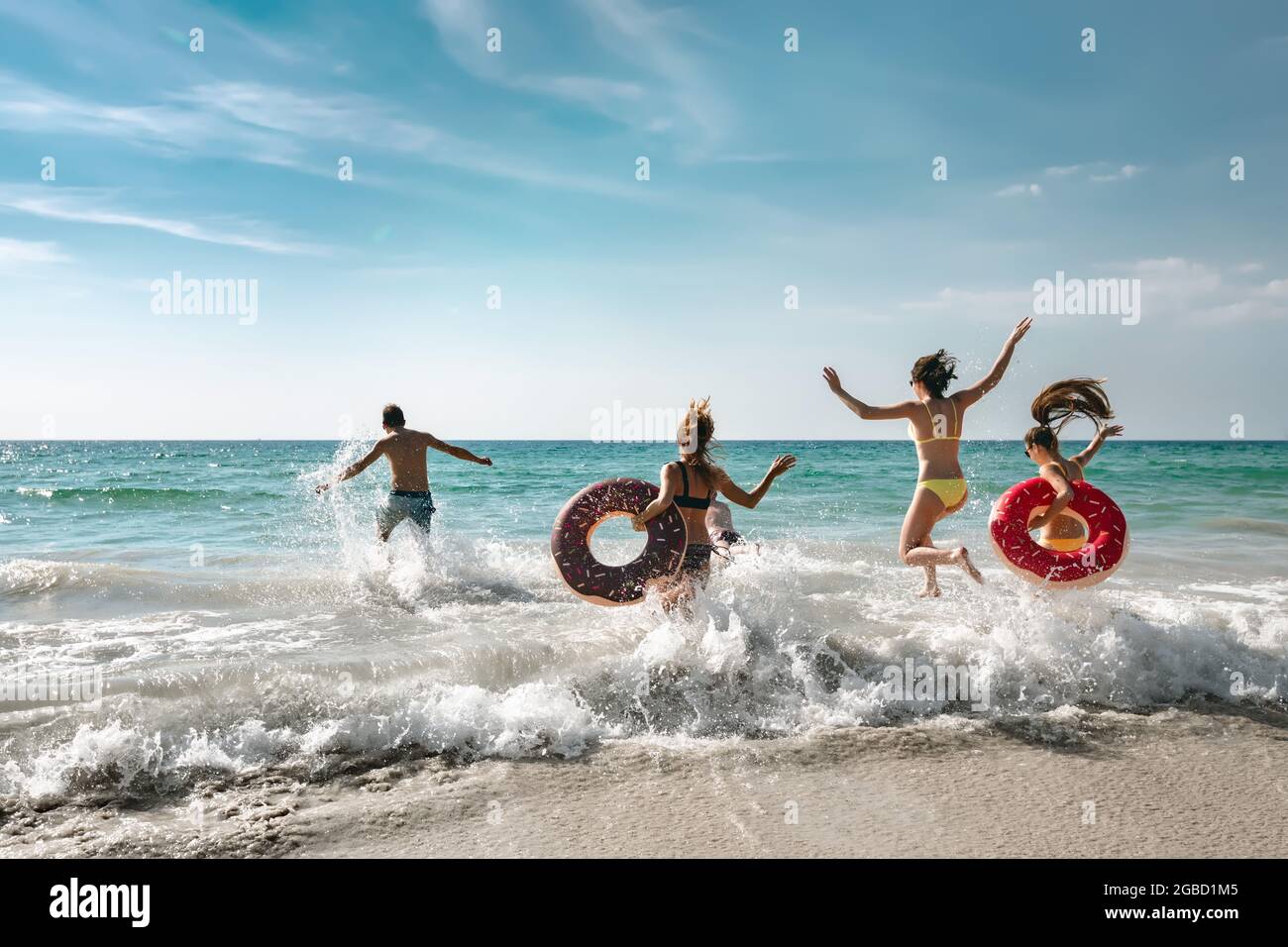 Fröhliche, fröhliche Freunde, die sich an sonnigen Tagen an einem schönen Strand ausruhen und mit aufblasbaren Ringen ins Meer laufen. Tropisches Urlaubskonzept Stockfoto