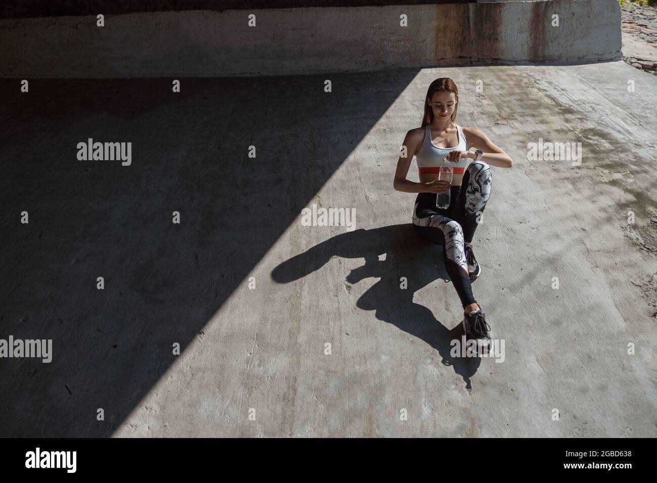 Frau in Sportkleidung und Turnschuhen öffnet eine Flasche Wasser auf Betonboden Stockfoto