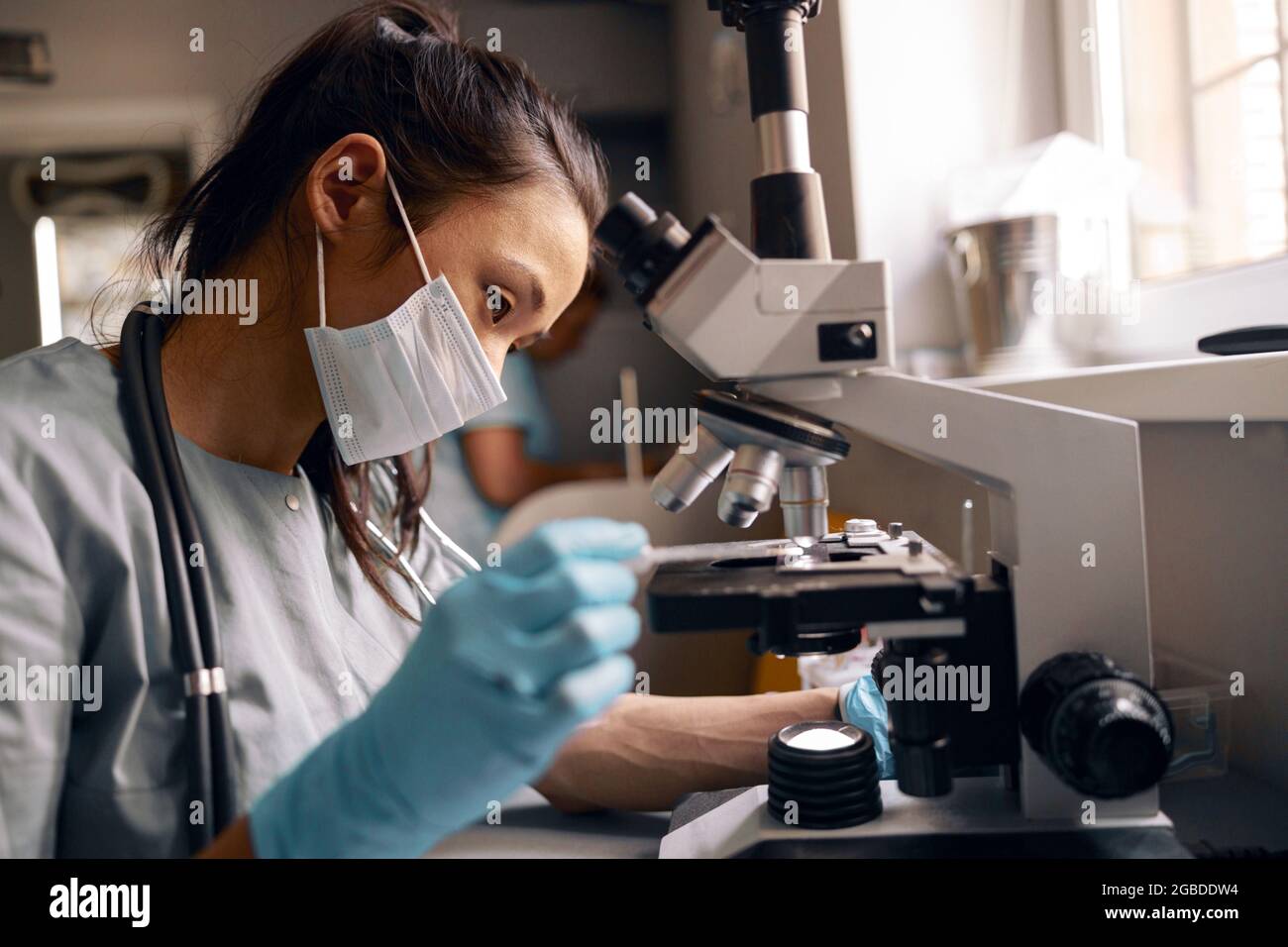 Asiatische weibliche Laborassistentin mit Maske bringt die Probe mit dem Mikroskop im Krankenhaus zur Forschung Stockfoto