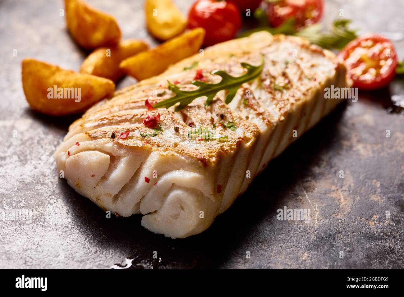 Appetitlich organischer Pollack-Fisch auf dunkler Oberfläche mit leckeren Kartoffelkeilen und Kirschtomaten Stockfoto