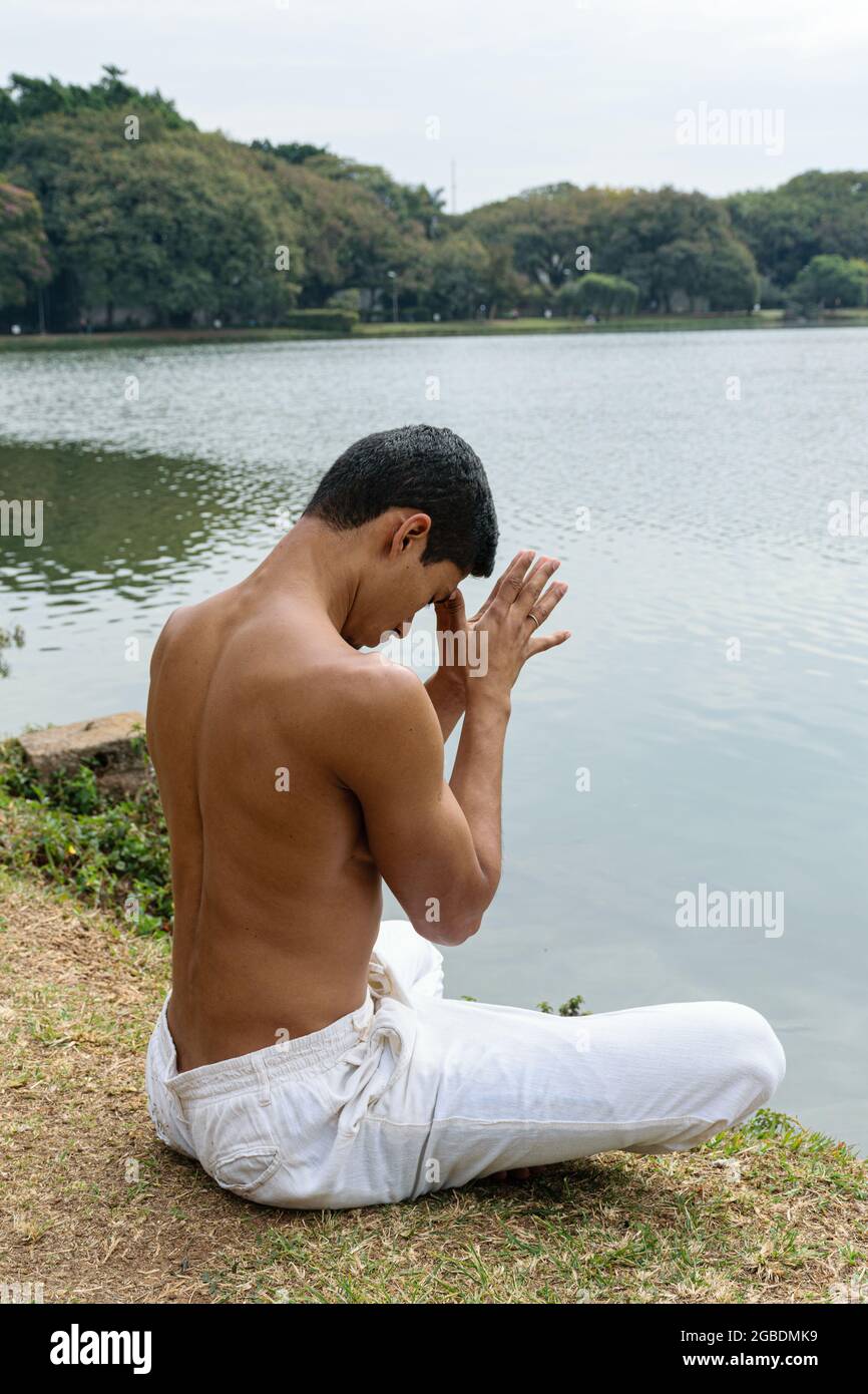 Brasilianischer junger Mann, der am Rande eines Sees sitzt, seine Hände gegenüber und die Daumen die Stirn berühren. Stockfoto