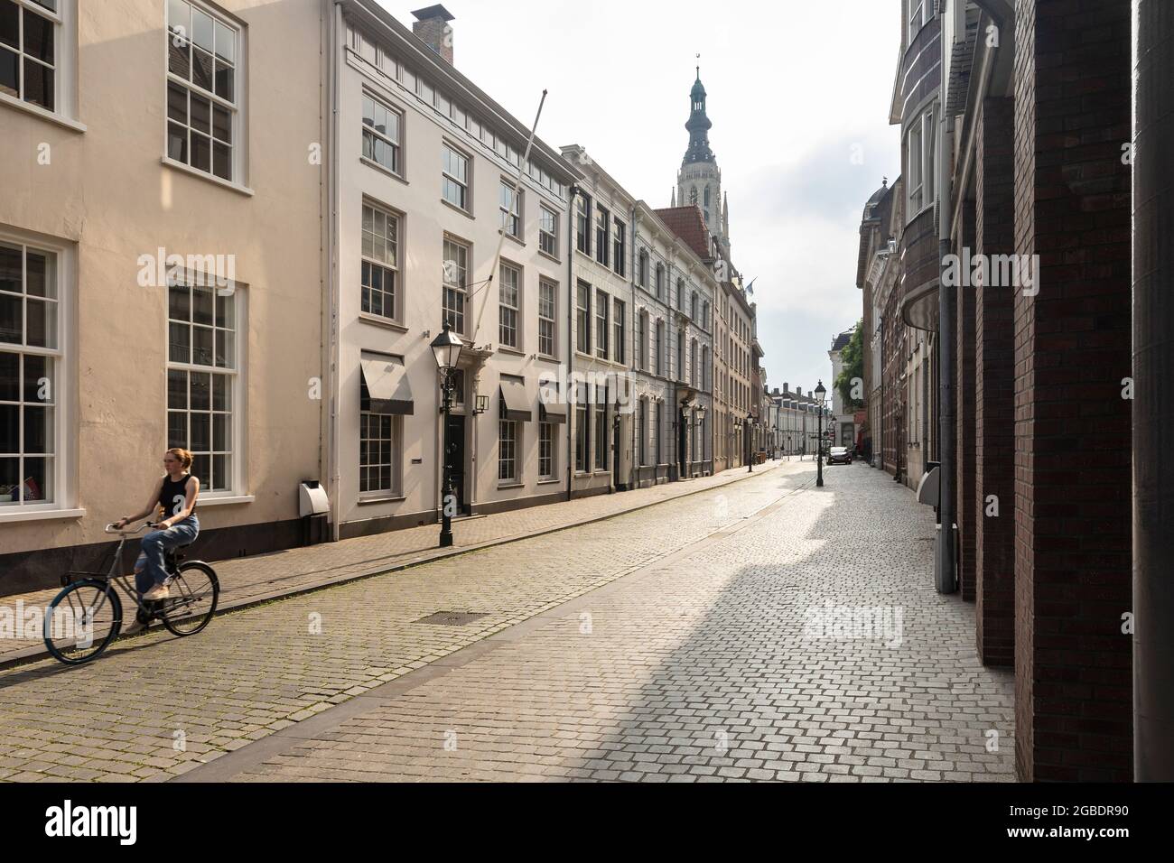 Breda, Niederlande 28. Juni 2021. Eine Straße im Stadtzentrum mit alten, hellen historischen Gebäuden, einem Bürgersteig, einer Kirche und einem Mädchen, das auf einem Su radeln muss Stockfoto