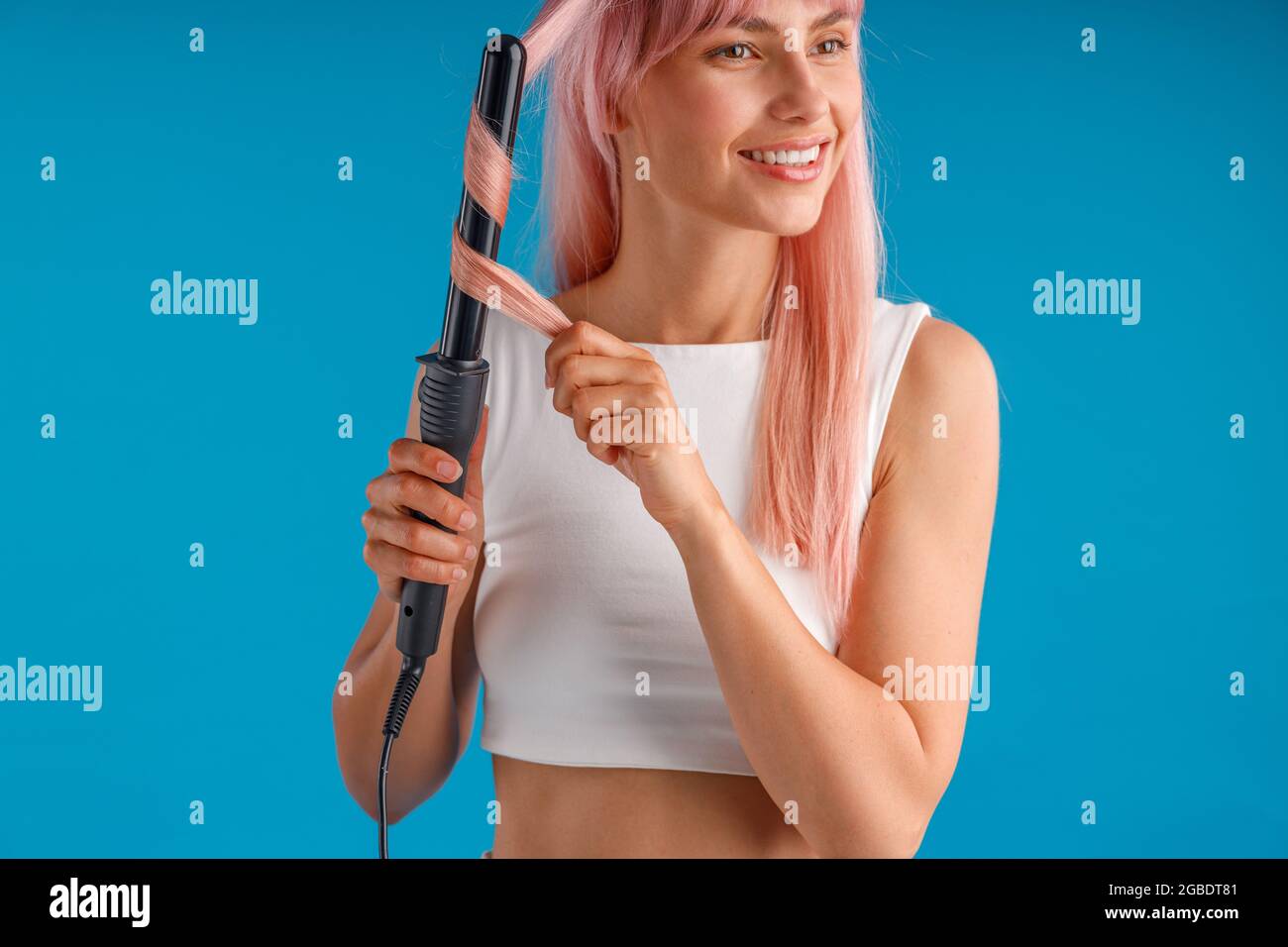 Schöne lächelnde Frau Styling rosa lange gerade Haare mit Lockenstab, isoliert stehend über blauen Studio-Hintergrund Stockfoto