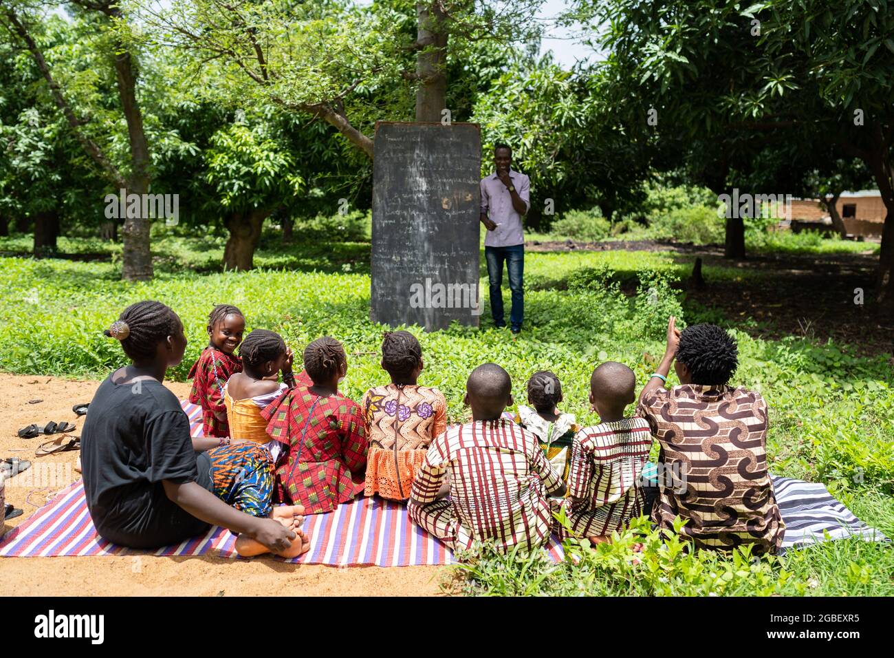 In diesem Bild hört eine Gruppe von Kindern, die auf einer Matte sitzen, ihrem Lehrer in einer Freiluftklasse in einer armen afrikanischen Dorfschule zu Stockfoto
