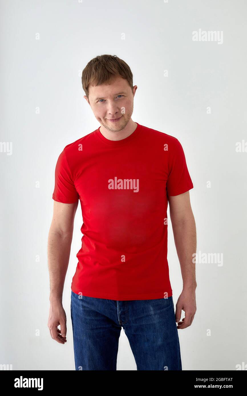 Positive junge männliche Modell in lässigem roten T-Shirt und Jeans Blick auf die Kamera, während sie vor weißem Hintergrund Stockfoto