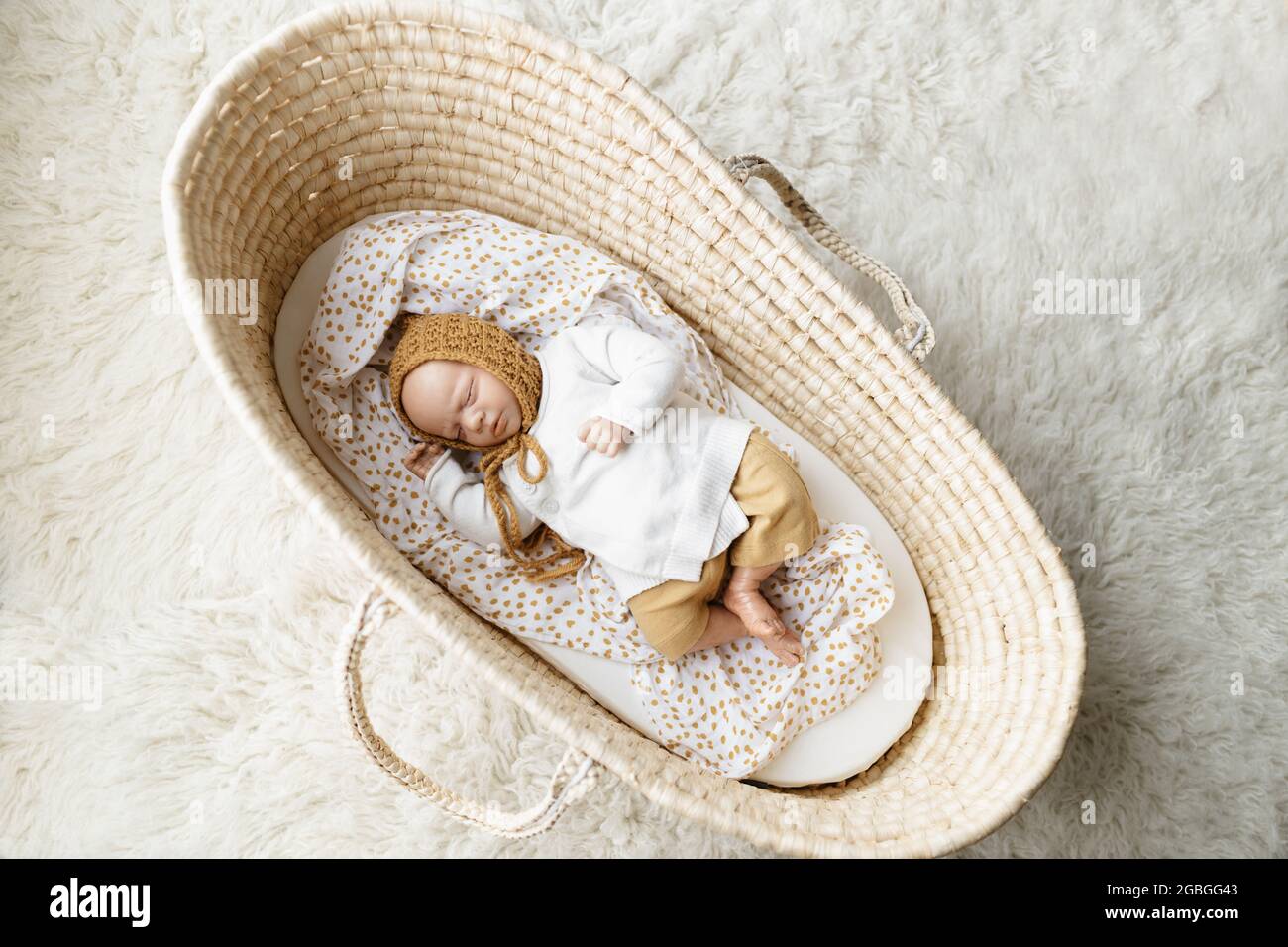 Ein neugeborenes Baby-Puppenspielzeug, gekleidet in einen senffarbenen Hut und eine weiße Strickjacke, die in einem cremefarbenen moses-Korb-Korb-Laderett liegt Stockfoto
