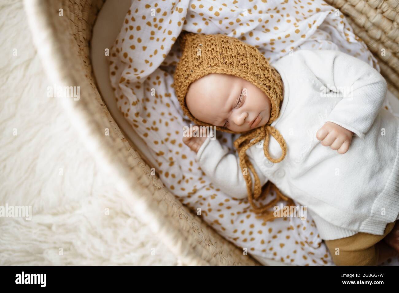 Ein neugeborenes Baby-Puppenspielzeug, gekleidet in einen senffarbenen Hut und eine weiße Strickjacke, die in einem cremefarbenen moses-Korb-Korb-Laderett liegt Stockfoto