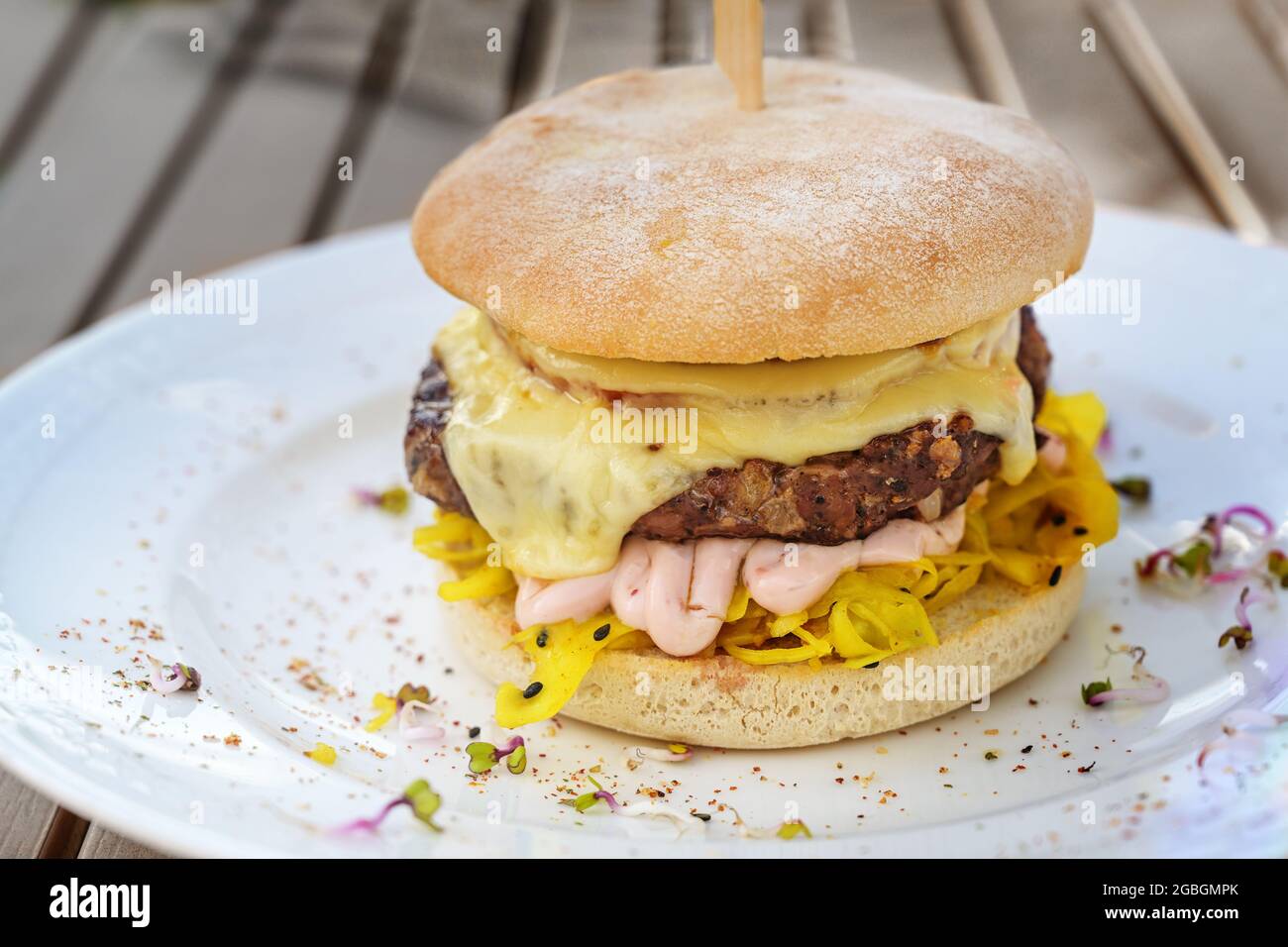 Burger mit einem groben Fleischpastete, Käse, Mayonnaise und Safrankohlsalat, garniert mit Gewürzen und Rettichsprossen auf einem weißen Teller und einem hölzernen Stockfoto