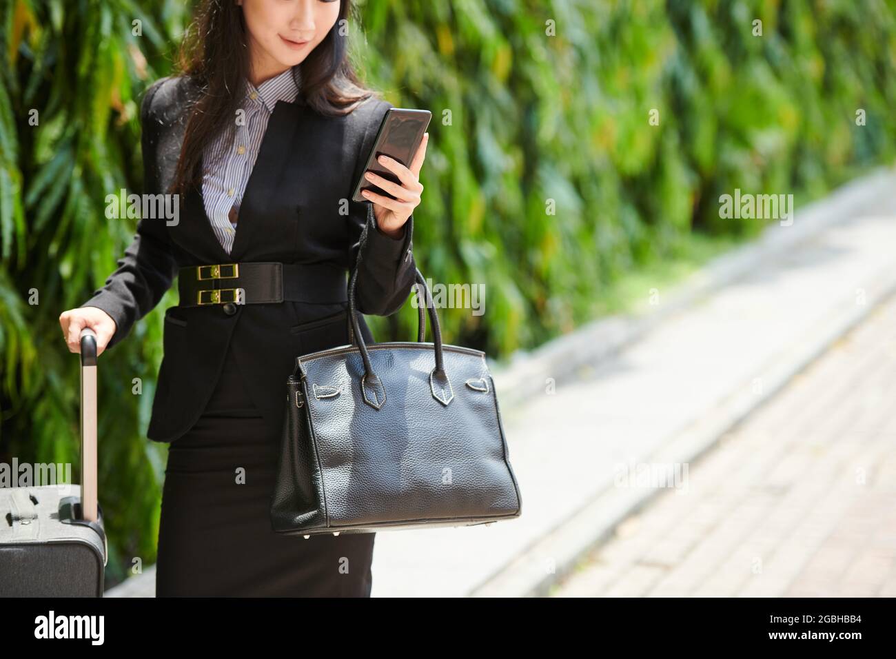 Inhalt junge asiatische Frau in formalwear, die Tasche und Koffer hält, während sie Informationen über den Flug am Telefon überprüft Stockfoto