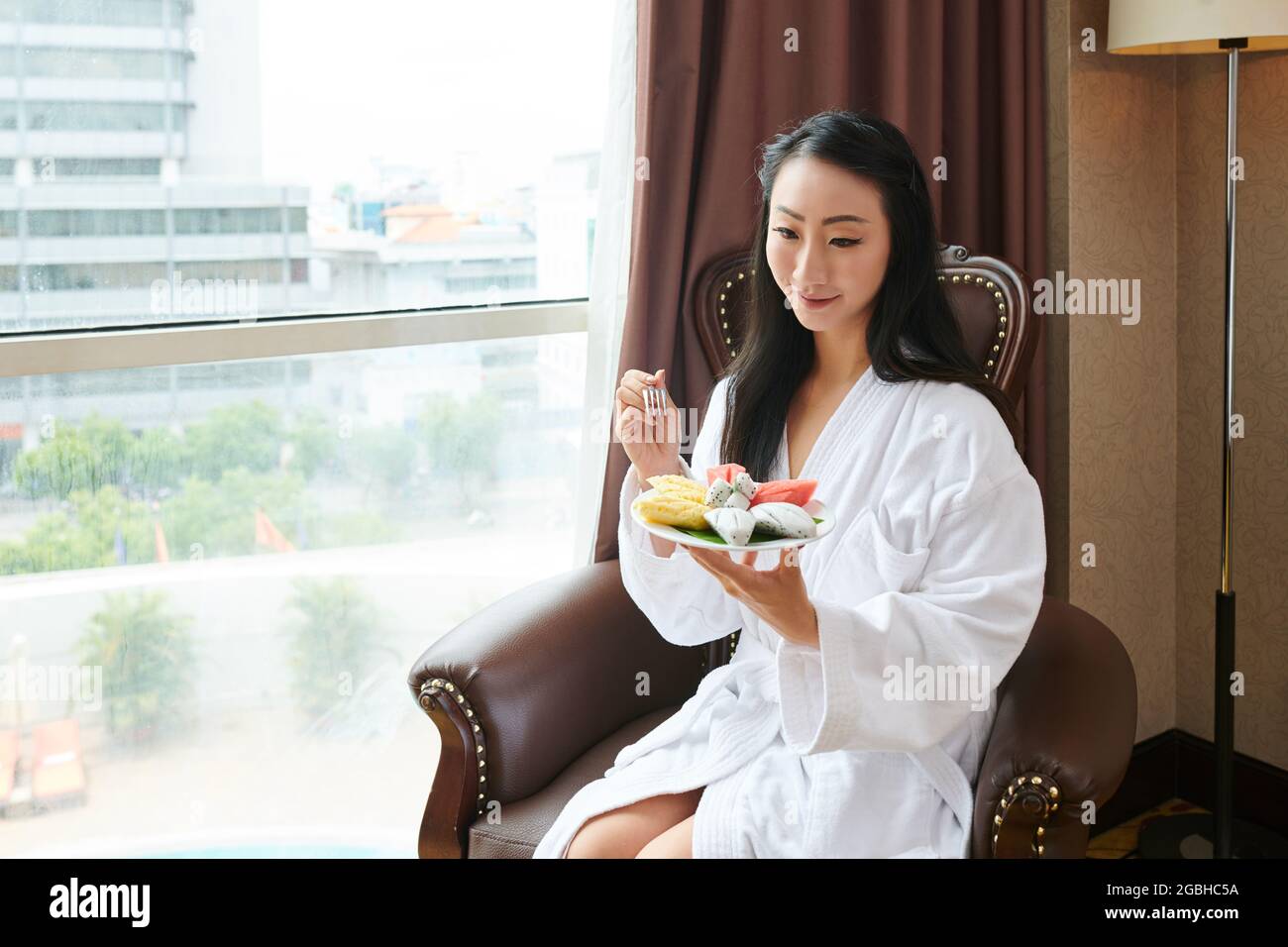 Attraktive junge asiatische Frau im weißen Hotelbademantel, die in einem Sessel am Fenster sitzt und tropische Früchte isst Stockfoto